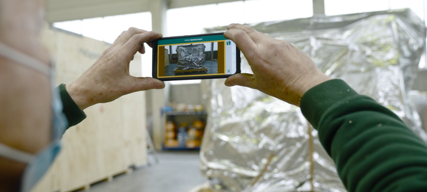 Hände halten ein Smartphone, das eine verpackte Ware in einer Lagerumgebung zeigt