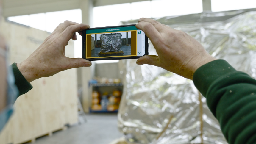 Hände halten ein Smartphone, das eine verpackte Ware in einer Lagerumgebung zeigt