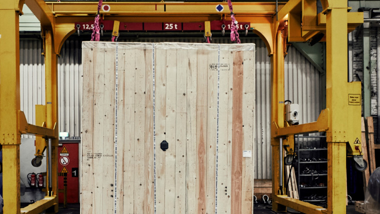 Große Holzkiste wird von gelbem Kran mit der Aufschrift Traglast 50 t in einer Lagerhalle gehoben