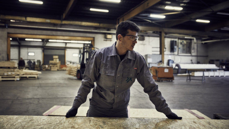 Ein Mitarbeiter mit Schutzbrille steht in einer Lagerhalle an einem Holzbrett