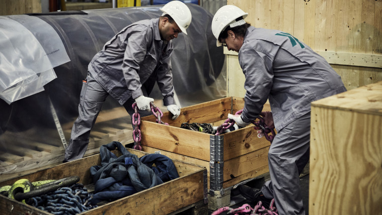 Zwei Personen in grauer Arbeitskleidung und Schutzhelmen heben gemeinsam eine Kette aus einem Holzkasten