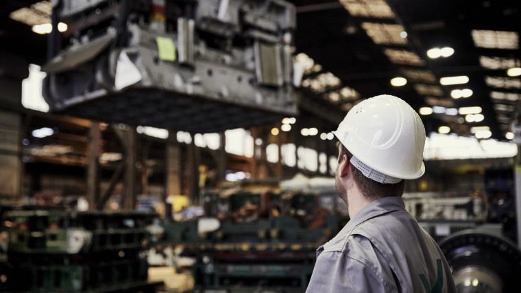 Ein Mann mit weißem Schutzhelm und grauer Arbeitskleidung blickt in einer großen Industriehalle auf eine hängende schwere Metallmaschine.