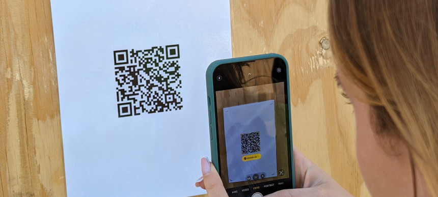 Frau hält Smartphone und scannt QR-Code auf weißem Blatt an Holzplatte
