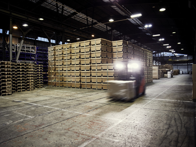 In einer dunklen Lagerhalle mit hochgestapelten Gütern, transportiert ein Gabelstapler eine Kiste.