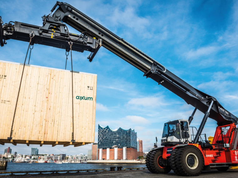 Ein Greifstapler hebt eine Groß0e AXXUM Exportverpackung aus Holz an. Im Hintergrund ist die Elbphilharmonie in Hamburg zu sehen.