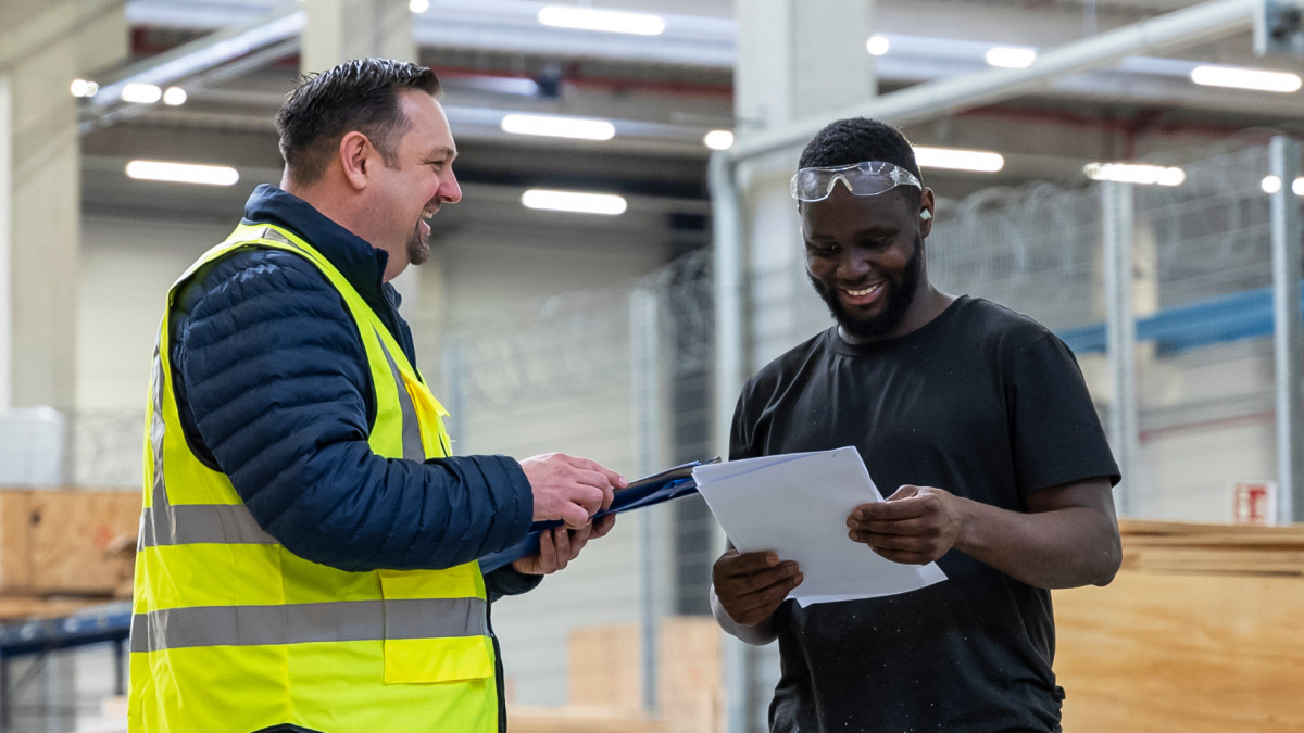 Zwei Mitarbeiter reden in einem Lager. Einer der Mitarbeiter trägt eine Warnweste, der andere hält eine Liste in der Hand