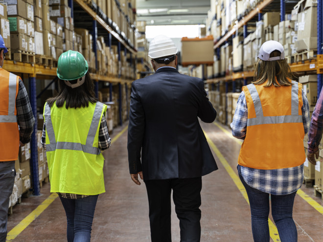 5 Personen, von den alle außer der Person in der Mitte eine Warnweste tragen, gehen durch einen Gang in einer Lagerhalle