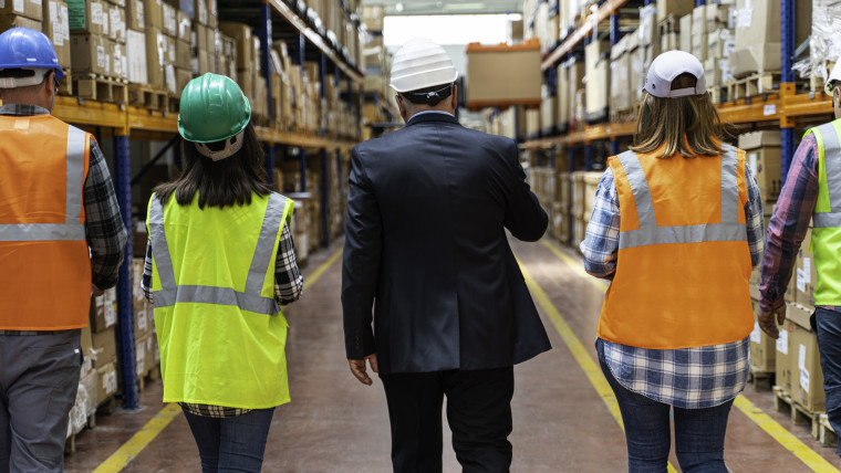 5 Personen, von den alle außer der Person in der Mitte eine Warnweste tragen, gehen durch einen Gang in einer Lagerhalle
