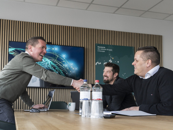 Drei Männer in einem Meetingraum. Der eine Mann steht und schüttelt einem anderen die Hand