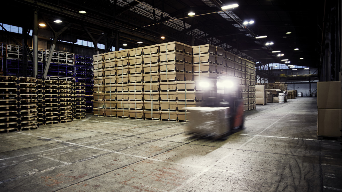 In einer dunklen Lagerhalle mit hochgestapelten Gütern, transportiert ein Gabelstapler eine Kiste.
