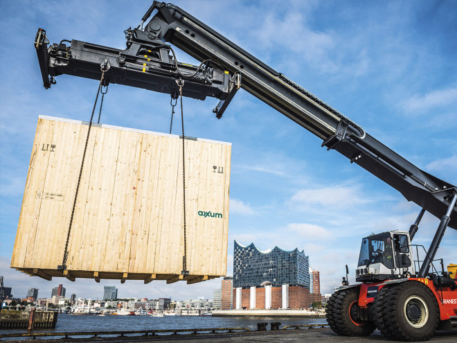 Ein mobiler Kran hebt eine Groß0e AXXUM Exportverpackung aus Holz an. Im Hintergrund ist die Elbphilharmonie in Hamburg zu sehen.