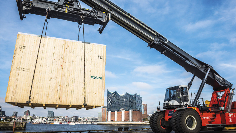 Ein mobiler Kran hebt eine Groß0e AXXUM Exportverpackung aus Holz an. Im Hintergrund ist die Elbphilharmonie in Hamburg zu sehen.