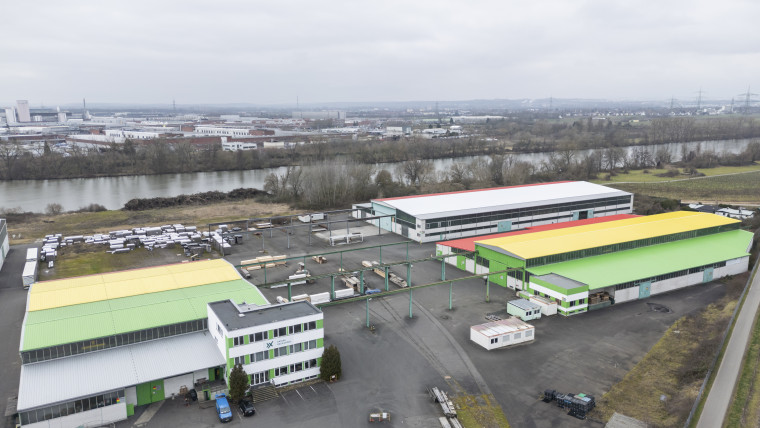 Luftaufnahme von dem Betriebsgelände von AXXUM Packaging Hessen mit mehreren großen Lagerhallen und einem Fluss im Hintergrund
