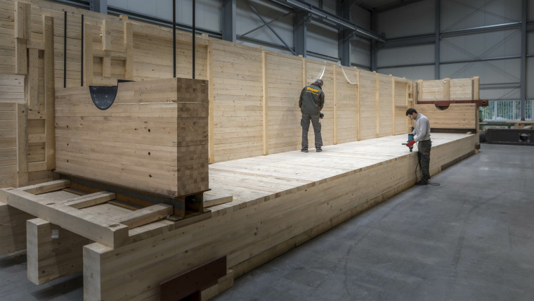 Zwei Männer arbeiten an einer großen Holzplattform in einer Halle mit hohen Wänden aus Holzpaneelen
