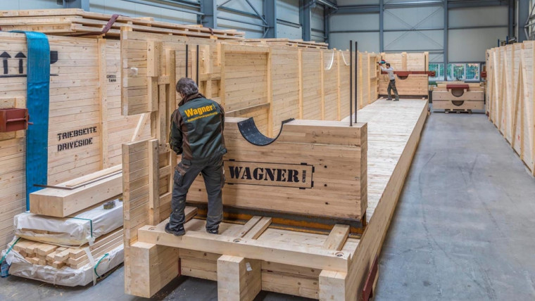Zwei Männer arbeiten an einer großen Holzplattform in einer Halle mit hohen Wänden aus Holzpaneelen