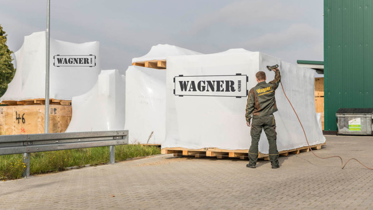 Mehrere große Transportkisten auf Paletten, die mit weißer Folie umwickelt sind, ein Mann in Arbeitskleidung mit Schriftzug Wagner steht daneben und hält ein Seil